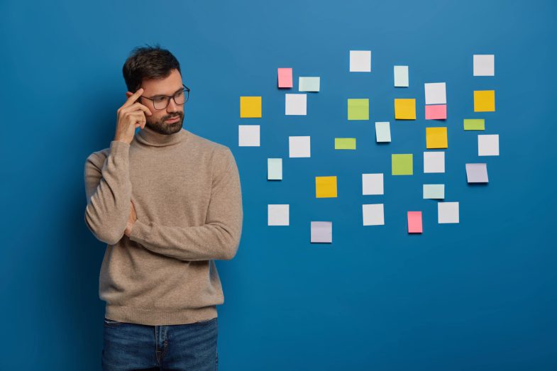 people-work-thoughts-concept-contemplative-bearded-guy-keeps-finger-temple-looks-pensively-aside-puts-colorful-sticky-notes-wall-min
